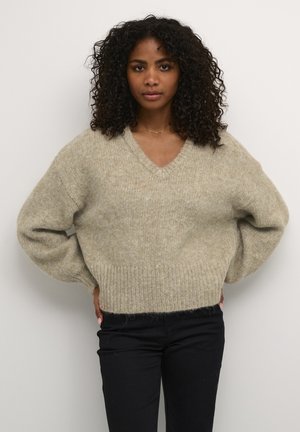 V-neck, oversized knit sweater in light beige, featuring a textured, soft fabric, ribbed cuffs, and hem. Paired with black pants.