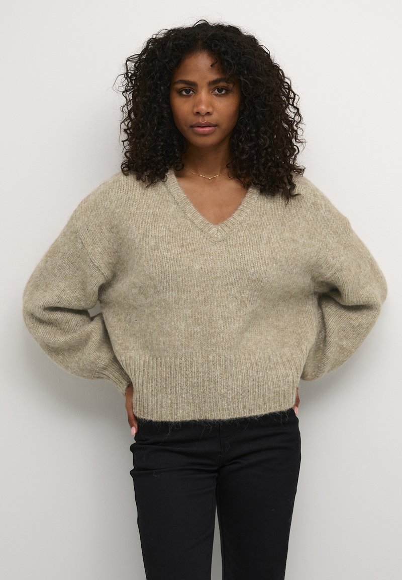Pull en maille oversize à col en V de couleur beige clair, présentant un tissu texturé et doux, avec des poignets et un ourlet côtelés. Assorti avec un pantalon noir.