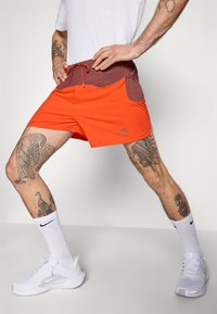 Mann trägt orangefarbene und braune Sportshorts, dehnt seine Beine, sichtbare Tattoos an Beinen und Armen, weiße Nike-Socken und Sneakers.