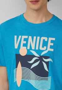 Blaues T-Shirt mit einem grafischen Design, das das Wort "VENEDIG", abstrakte Formen und wellenförmige Linien in Marine- und hellen Farbtönen zeigt. Aus Baumwollmaterial.