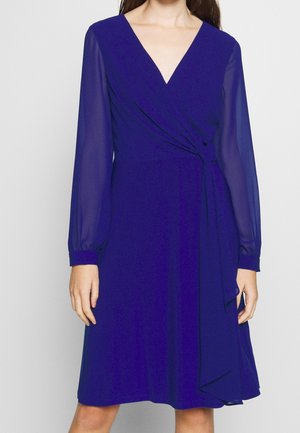 Mujer con vestido cruzado azul real hasta la rodilla, con mangas largas transparentes y escote en V profundo.