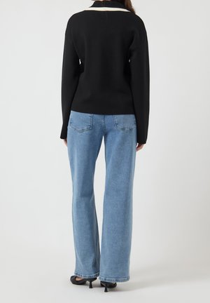 Femme portant un jean bleu clair droit, un pull noir avec un col blanc et des chaussures noires à talons, debout de dos.