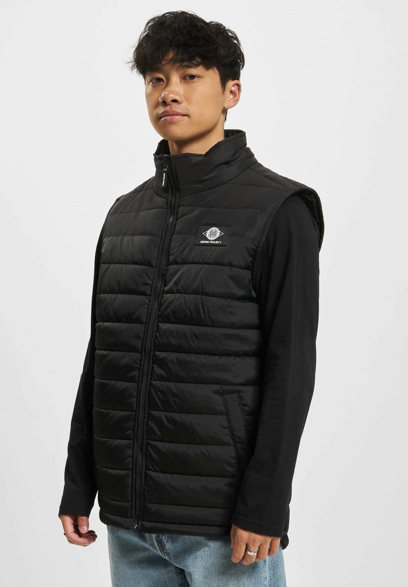 Svart polstret vest med glidelås, høy krage og logopatch. Har en glatt tekstur og sidelommer, bæres over en langermet skjorte.