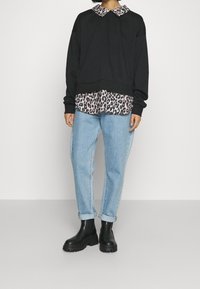 Person som bär en svart tröja över en leopardmönstrad skjorta, ljusblå upprullade jeans och svarta kraftiga ankelskor, stående mot en enkel bakgrund.