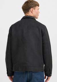 Chaqueta de ante negra con cuello, corte recto y costuras visibles. Presenta puños abotonados y se combina con jeans de denim azul.