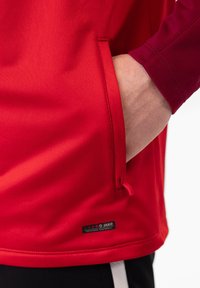 Rotes Sportshirt mit strukturiertem Finish, Seitentasche und kontrastierenden kastanienbraunen Ärmelbündchen; mit einem JAKO-Label am Saum versehen.