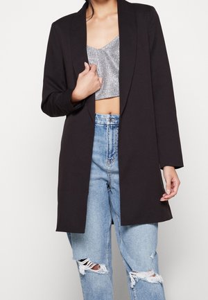 Sort blazer med åben frontdesign, lange ærmer, glat tekstur, båret over en sølvfarvet mønstret crop top og lyseblå slidte jeans.