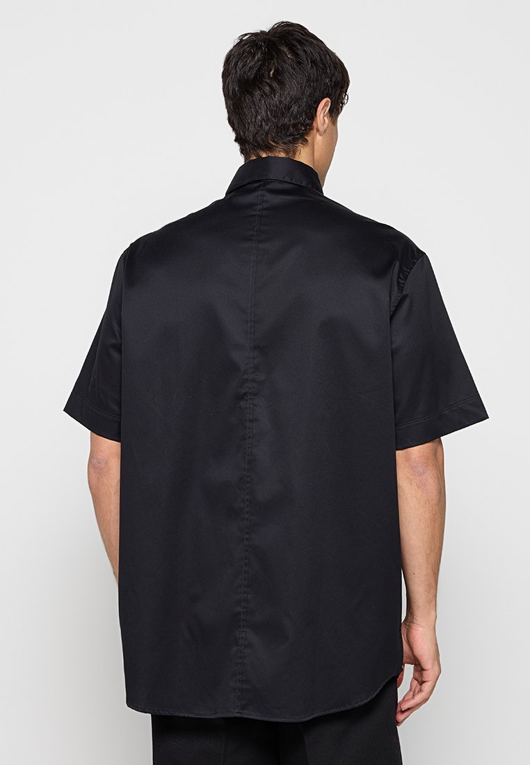Hombre con camisa negra de manga corta y cuello, visto desde atrás contra un fondo claro y liso.