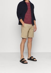 Mörkblå knappad jacka, vinröd texturerad t-shirt, khakishorts och svarta sandaler med två remmar. Fokusera på avslappnad, lager-på-lager stil.
