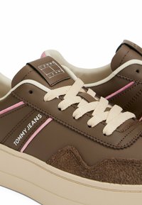 Baskets marron composées de cuir et de daim, avec des semelles crème, des accents roses et des lacets épais. Présente le nom de la marque "TOMMY JEANS" sur le côté.