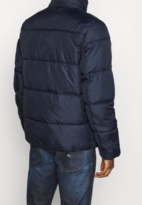 Granatowa kurtka puchowa z poziomym przeszyciem, wysokim kołnierzem i gładką, błyszczącą teksturą. Noszona z niebieskimi dżinsami, widok z tyłu.