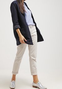 Cardigan navy a trama, camicia azzurra chiara, pantaloni beige chiari arrotolati e mocassini argentati indossati da una persona, che mostrano un abbigliamento casual a strati.