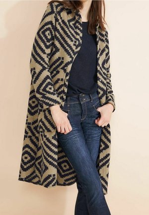 Kobieta ubrana w beżowy płaszcz w czarne geometryczne wzory, na czarnym topie i ciemnoniebieskich dżinsach, stojąca z rękami w kieszeniach.