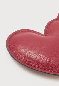 Κόκκινο δερμάτινο αξεσουάρ σε σχήμα καρδιάς με εμφανείς ραφές και ανάγλυφο το όνομα της μάρκας "FURLA" σε λευκό φόντο.