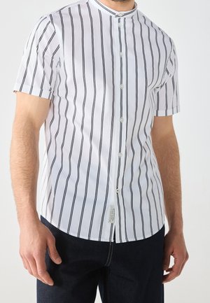 Homme portant une chemise blanche à manches courtes avec des rayures verticales noires et un pantalon foncé, debout avec les bras détendus.