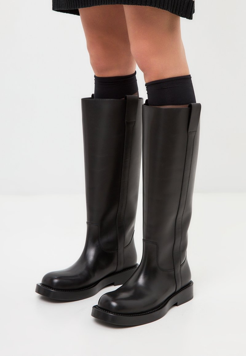 Bottes noires hautes jusqu'au genou en cuir lisse, dotées d'un bout arrondi et d'une semelle en caoutchouc plate. Design minimaliste avec des lignes épurées et sans ornements.