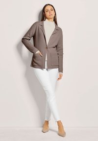 Hellbraune Strickjacke mit einem einzelnen Knopf, gerippten Ärmeln und zwei Fronttaschen, kombiniert mit einem cremefarbenen Rollkragenpullover und weißen Hosen.