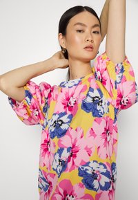 Vestido floral com grandes flores rosas e azuis sobre um fundo amarelo. As mangas bufantes e a gola redonda adicionam textura e forma.