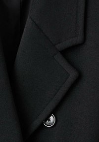 Czarna wełniana tkanina z ostrym klapem i wyraźnym przeszyciem. Ozdobiona okrągłym czarnym guzikiem z trzema otworami.