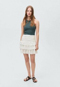 Geribbelde donkergroene tanktop gecombineerd met een wit, gelaagd minirok met plooien. Zwarte platte sandalen maken de outfit compleet.