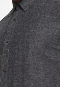 Camisa abotonada en espiga gris con cuello estándar, sutil textura y botones negros de plástico a lo largo de la tapeta delantera.