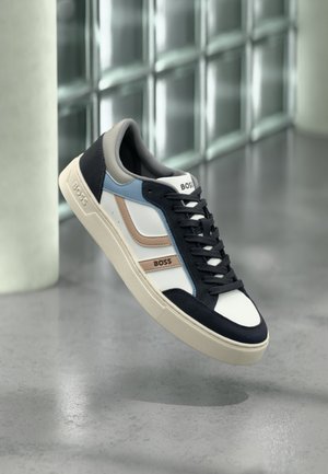 Sneaker BOSS bianco, blu navy, blu e beige con lacci neri, sospesa sopra un pavimento in cemento levigato vicino a una parete di blocchi di vetro.