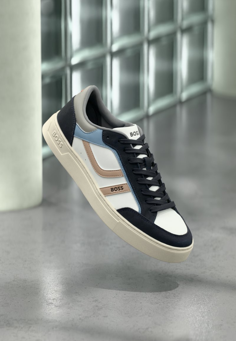 Baskets BOSS blanches, bleu marine, bleu et beige avec des lacets noirs, flottant au-dessus d'un sol en béton poli près d'un mur en parpaings de verre.