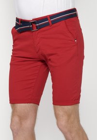 Shorts en coton rouges avec un ourlet roulé, dotés d'une ceinture bleu marine avec des rayures rouges et blanches. Poche latérale et un détail de marquage minimal.