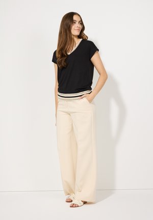 Haut noir à manches courtes avec un ourlet rayé crème, associé à un pantalon large crème et des sandales simples à lanières sur fond blanc.