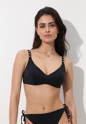 Donna con capelli lunghi e scuri che indossa un bikini nero con spalline accentate in oro, in piedi davanti a uno sfondo bianco uniforme.