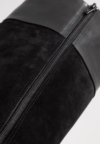 Bottine en daim noir et cuir avec une fermeture éclair visible et des détails cousus, mettant en valeur une partie supérieure lisse et une section inférieure texturée.