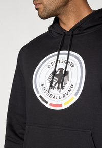 Fekete kapucnis pulóver, amelyen egy kiemelkedő, kerek logó látható, benne egy stilizált sas, a "Deutscher Fussball-Bund" felirattal és élénk színes keretekkel piros, fekete és sárga színekben.