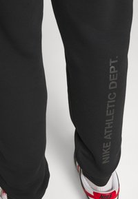 Svarta träningsbyxor med en slät yta, som har en vertikal grå logotyp med texten "NIKE ATHLETIC DEPT." som visas längs sidan.