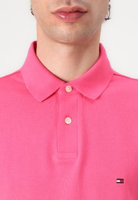Das rosa Poloshirt besteht aus einem strukturierten Stoff, hat einen umgeschlagenen Kragen und drei Knöpfe am Hals. Ein kleines Logo befindet sich unten links.