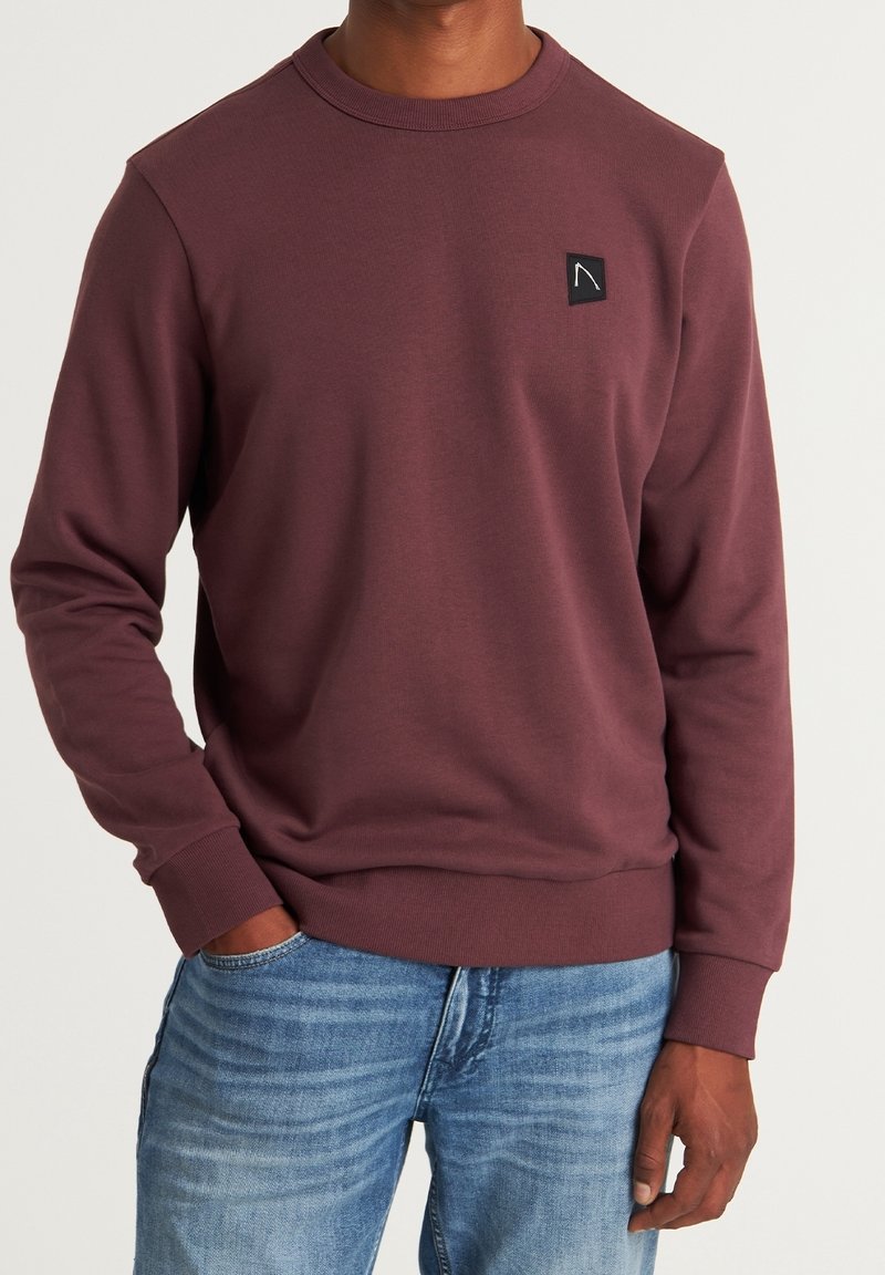Homme portant un sweat-shirt à manches longues bordeaux avec un petit patch logo noir sur la poitrine et un jean bleu, main dans la poche.