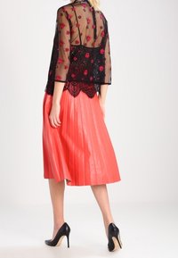 Top in pizzo nero con ricami floreali e maniche trasparenti, abbinato a una gonna a pieghe color corallo. Scarpe alte nere completano il look.