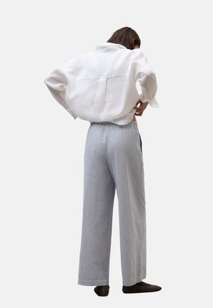 Mujer de pie con las manos en las caderas, vistiendo una camisa blanca holgada, pantalones anchos de tiro alto grises y zapatos oscuros sin cordones.