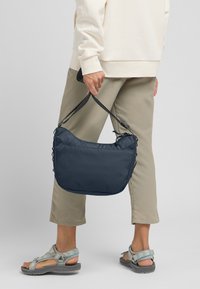 Borsa a spalla blu navy realizzata in tessuto testurizzato, caratterizzata da una forma morbida e arrotondata con una tracolla nera. Include chiusura con coulisse e dettagli laterali.