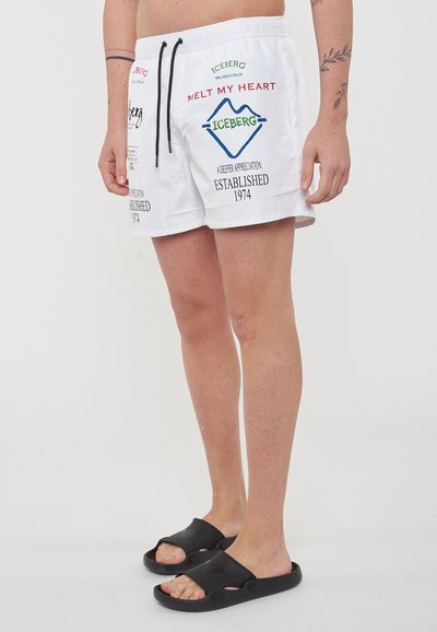 Pantaloni da bagno bianchi con vita regolabile, caratterizzati da testo e loghi colorati; indossati con sandali neri su uno sfondo semplice.