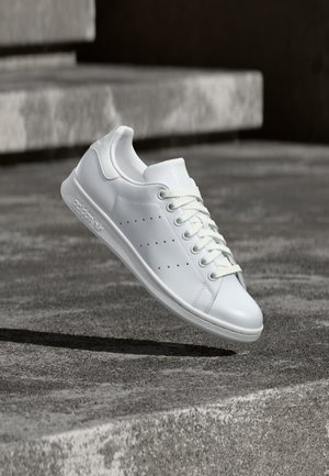 Hvide lavtop lædersneakers med snørebånd og perforerede sidelister, svævende over teksturerede betontrapper i blødt naturligt lys.