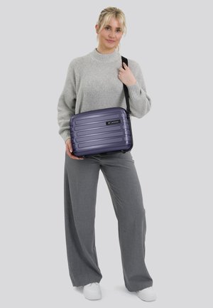 Borsa a tracolla viola con guscio rigido, striature orizzontali, dettagli neri e tracolla regolabile, tenuta da una persona in un abbigliamento grigio.
