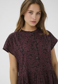 Robe à motifs floraux en bordeaux profond et noir. Dotée de manches courtes, d'un col rond et d'une patte de boutonnage sur le devant.