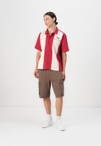 Rød og kremfarge kortermet skjorte med frontlukking og logo. Brune cargo shorts med sidelommer. Modellen har på seg hvite sneakers.