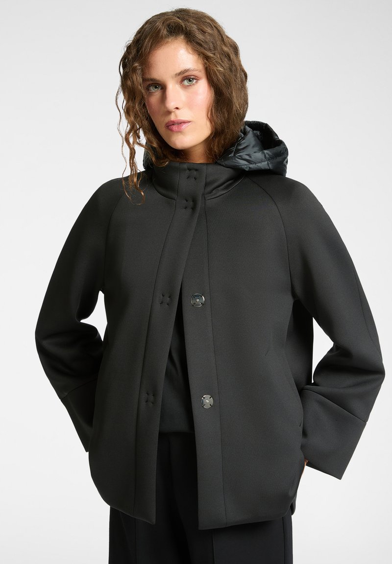 Czarna kurtka z wysokim kołnierzem i puchowym kapturem. Posiada metaliczne guziki, gładką fakturę oraz luźny krój z szerokimi rękawami.