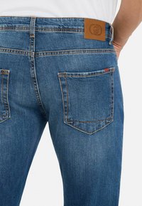 Jeans de mezclilla en azul medio con una textura suave. Presenta dos bolsillos traseros, costuras en contraste y una etiqueta de cuero marrón en la cintura.