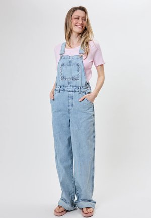 Lächelnde Frau mit langen blonden Haaren, die hellblauen Jeans-Latzhose, ein pinkes kurzärmeliges Oberteil und pinke Flip-Flops trägt, steht mit den Händen in den Taschen.