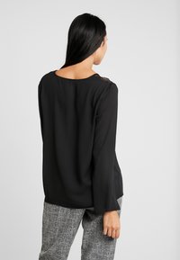 Czarna bluzka z długim rękawem, okrągłym dekoltem i koronkowymi zdobieniami na ramionach, zestawiona z szarymi spodniami o fakturze.