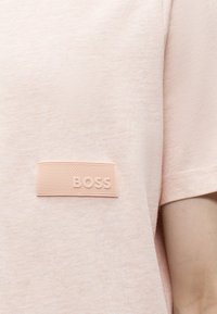 Chemise rose clair à manches courtes avec un écusson rectangulaire texturé portant l'inscription « BOSS » sur la poitrine, portée par une personne avec un bras visible.