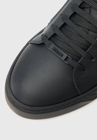 Tenis cu piele neagră, cu vârf rotund, suprafață texturată și șireturi groase negre. Are un mic logo aproape de zona șiretului.