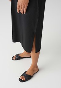 Zwarte mouwloze jurk met een zijscheur, gedragen met zwarte instapsandalen met kruislings straps en vierkante neuzen. Glad stoftextuur.